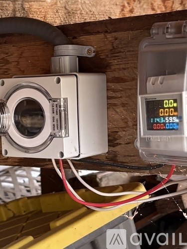 A digital device with a white body and a silver lens is connected to a yellow structure with a red and black wire.