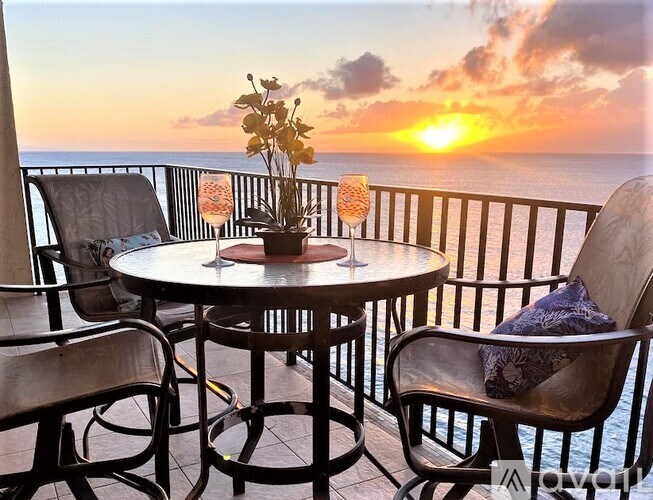 A table with chairs and a vase of flowers on a balcony overlooking the ocean at sunset.