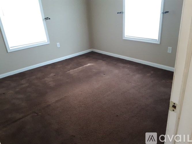 A room with brown carpet and two windows.