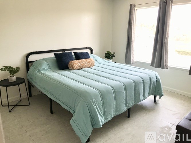 A bed with a blue comforter and two pillows sits in a room with a window and a plant.