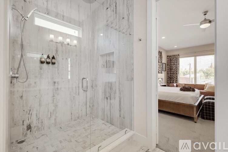 A bathroom with a walk-in shower and a bedroom with a bed and a window.