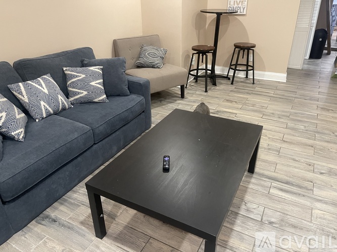 A black coffee table with a remote on it sits in front of a grey couch with grey and white pillows.