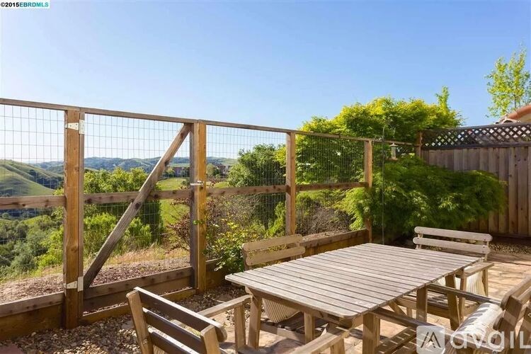 A wooden table and chairs are set on a patio with a view of the hills.