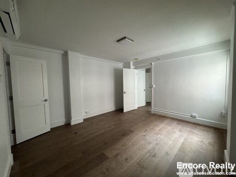 A room with white walls and wooden flooring, with a door on the left and a window on the right.