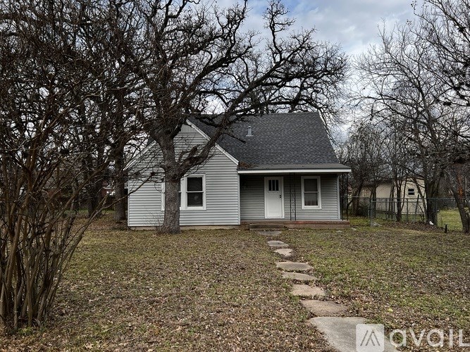 A small house with a grey roof and a white door is surrounded by trees.