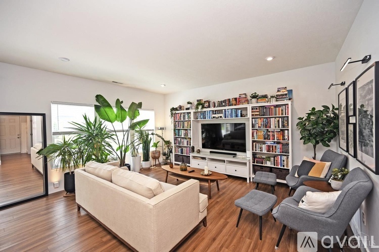 A living room with a white couch, a grey armchair, a wooden coffee table, a bookshelf filled with books, and a large window.