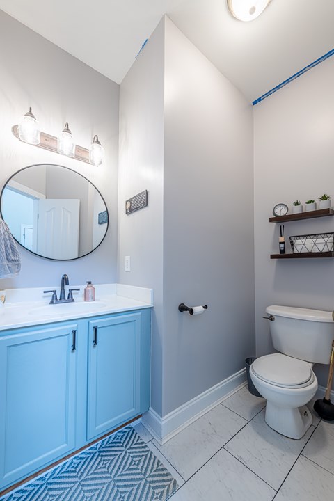 A white toilet sits in a bathroom with blue cabinets and a round mirror.