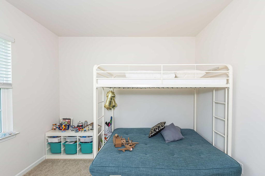 A white bunk bed with a blue mattress and pillow is in a room with a window and a shelf with toys.