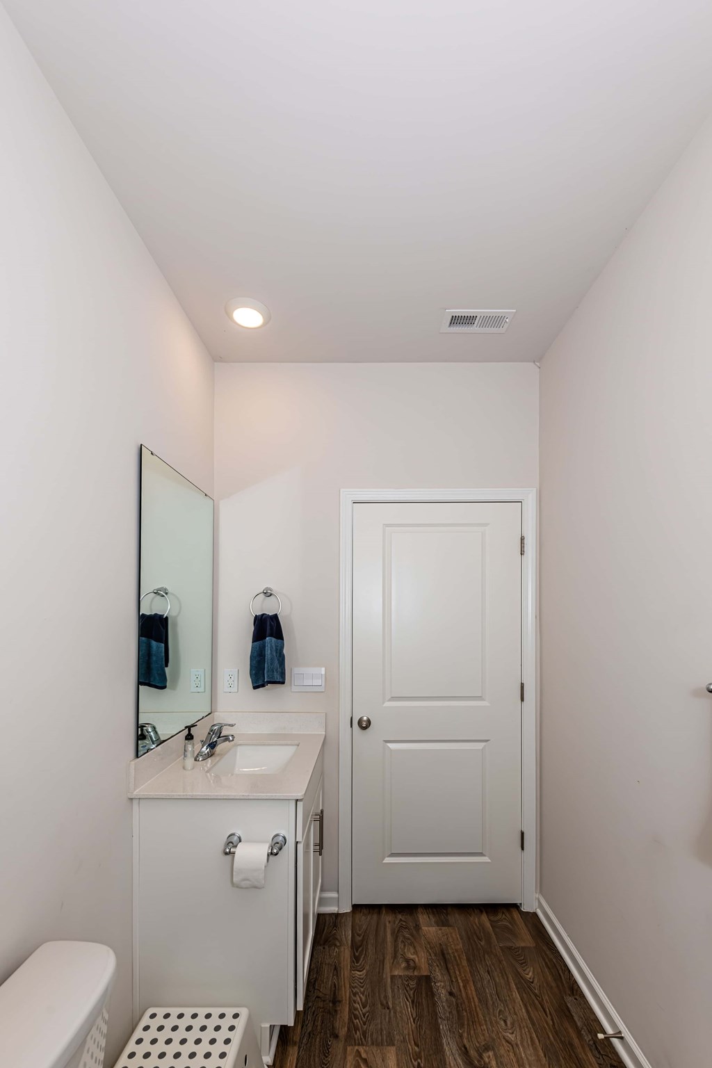 A white bathroom with a toilet, sink, mirror, and wooden floors.