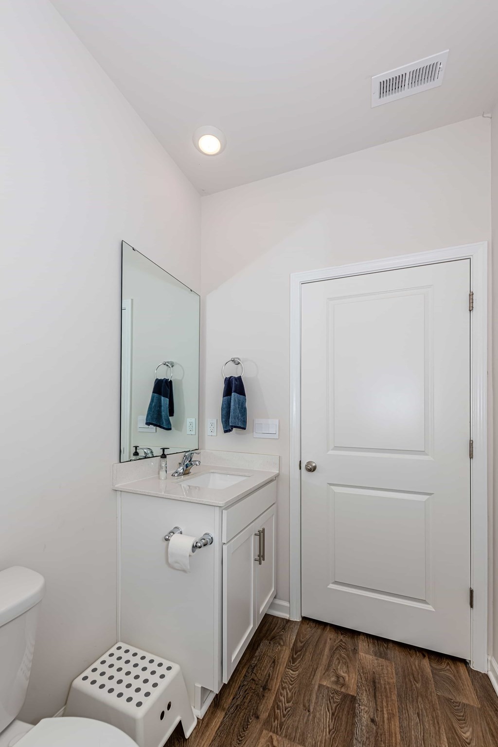 A white bathroom with a toilet, sink, mirror, and wooden floors.