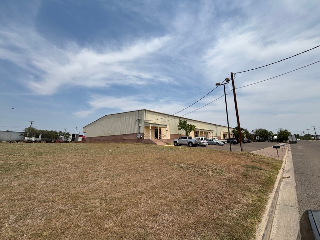 A large building with a grey roof is situated in a grassy area with a road in front of it.