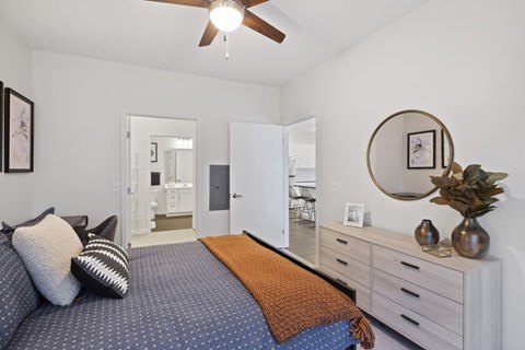 Fridley, MN, Luxury Apartments Near Mercy Hospital - Axle Apartments - Bright Bedroom With Ceiling Fan, Plush Bedding, Light Wood Dresser, Round Mirror, Home Decor, and Ensuite Bath Entry.