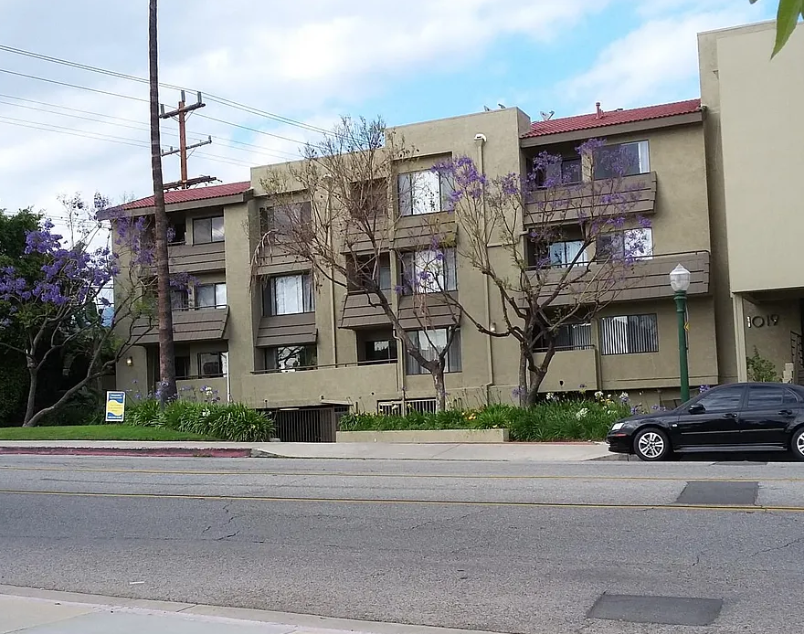 A black car is parked on the side of a street in front of a building with a tree in front of it.