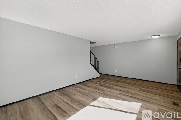 A room with wooden flooring and a white wall with a staircase in the background.