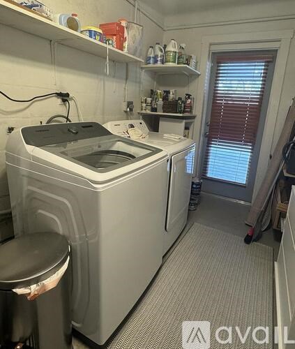 A small, white, industrial-sized washing machine sits in a small room with a shelf above it.