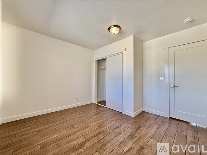 A room with wooden flooring and white walls, with a door on the right and a window on the left.