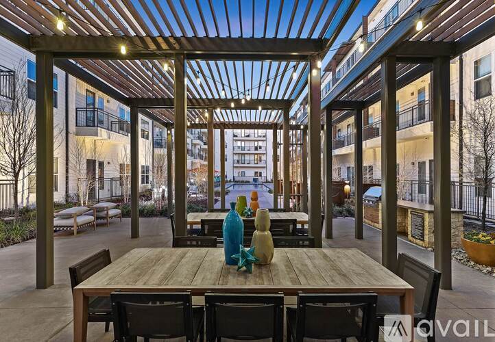 A wooden table with chairs and a vase on it is in the foreground of an outdoor seating area with a glass roof and buildings in the background.