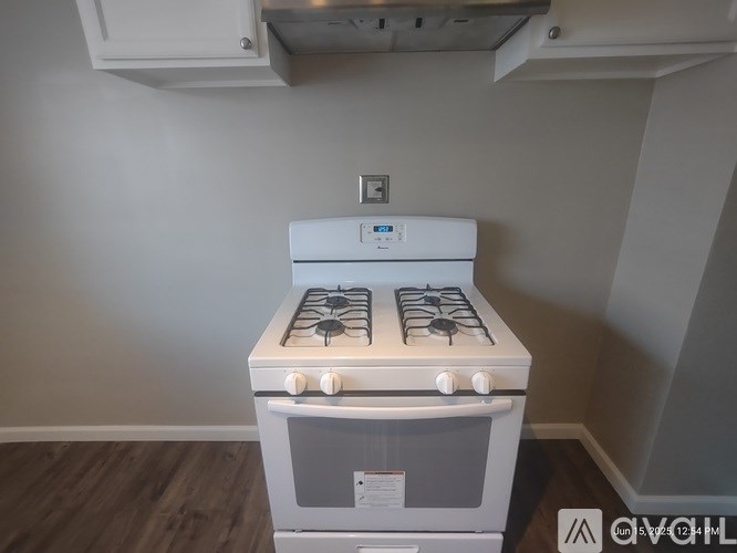 A white gas stove with a grey oven is in a room with a white wall and a wooden floor.