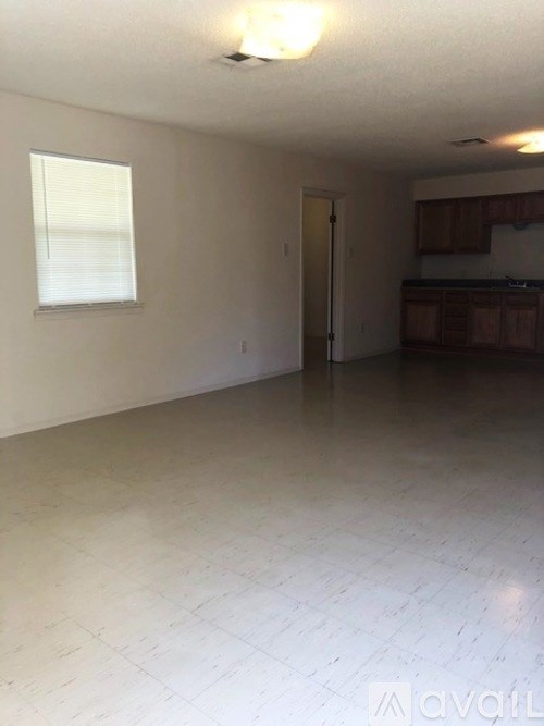 A large empty room with a window and a door leading to a kitchen.