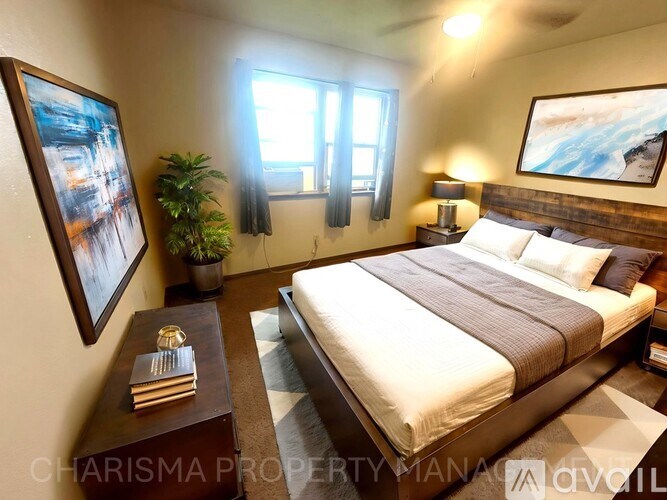 A well-lit bedroom with a large bed, a side table with a plant, and two framed pictures on the wall.