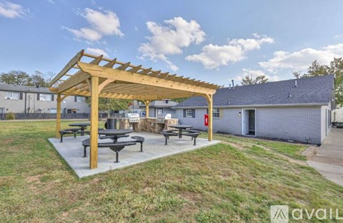 A wooden pergola with benches is situated in a grassy area with a building in the background.