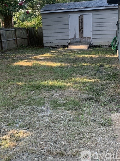 A small, dilapidated building sits in a yard with a fence and a hose nearby.