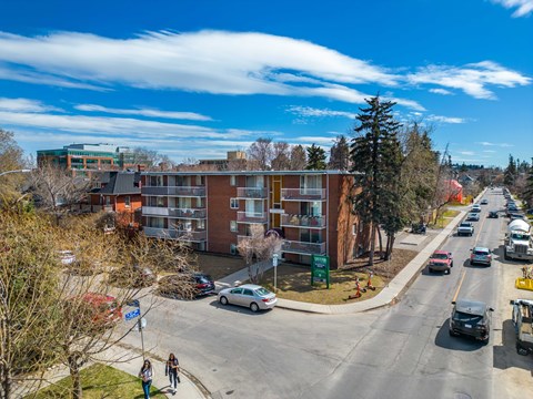 A street view of a residential area with cars parked on the side of the road and people walking on the sidewalk.