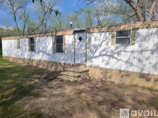 A white building with a brown roof and a door is surrounded by grass and trees.