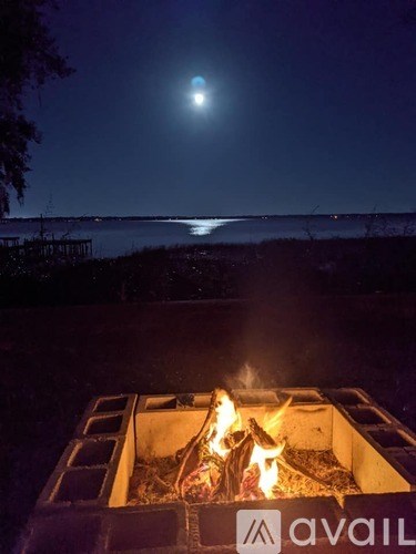 A campfire burns in a fire pit with a full moon rising over a body of water.