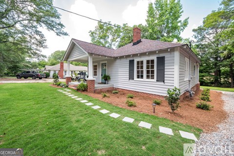 A small white house with a red brick chimney is surrounded by a green lawn and a gravel path.