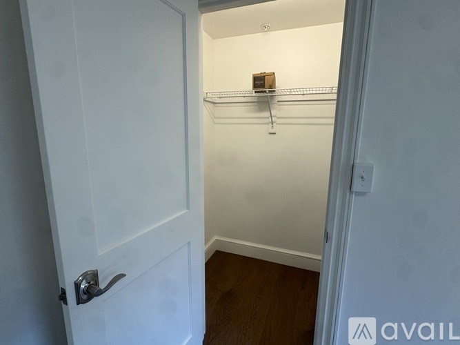 A white door with a silver handle is open to a hallway with a white wall and a brown floor.