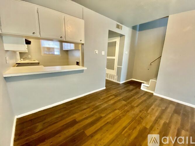 A room with wooden floors and white walls, with a kitchen area visible in the background.