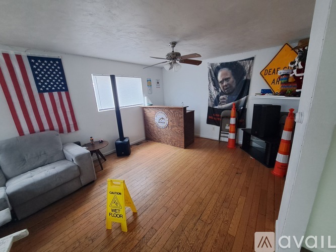 A living room with a grey couch, a wooden coffee table, a yellow caution sign on the floor, and a poster of a man's face on the wall.