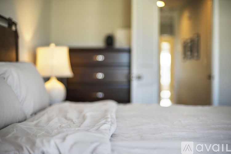 A bed with white sheets and a white pillow is in the foreground with a lamp and a dresser in the background.