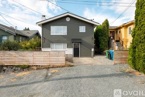 A grey house with a black door and windows is surrounded by a gravel driveway and a wooden fence.