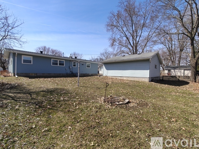 A blue house with a white garage is situated in a grassy area with trees in the background.