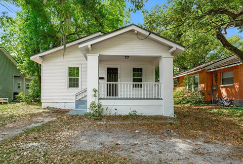 A small white house with a porch is surrounded by trees.