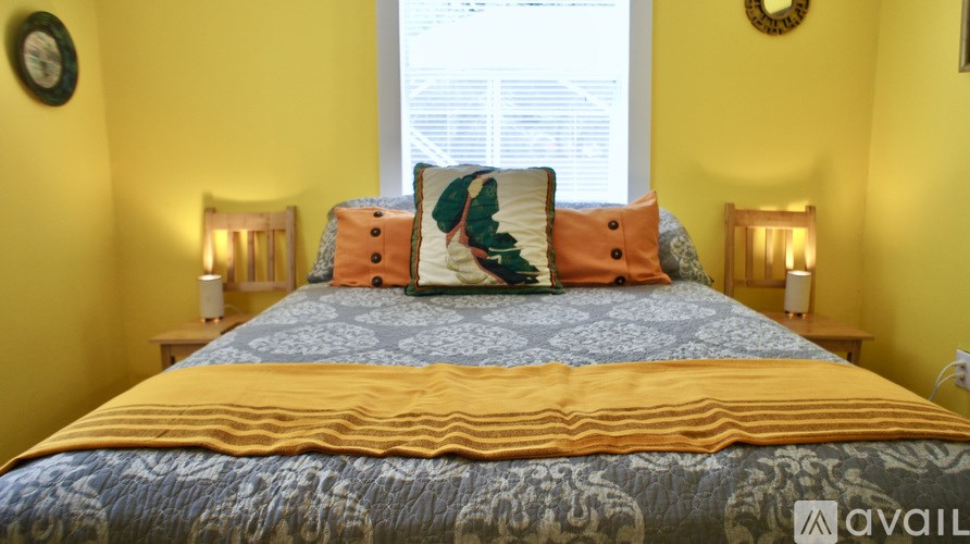 A bed with a yellow comforter and a blue and orange pillow is in the foreground of a room with yellow walls.