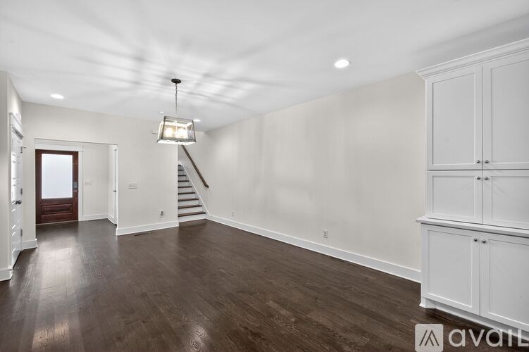 A spacious room with wooden floors and white walls, featuring a staircase and a chandelier.