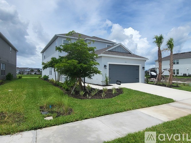 A house with a white exterior and a grey roof is surrounded by a well-maintained lawn and a small garden with a palm tree.