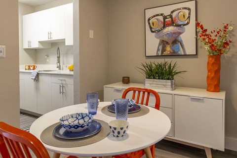 A dining room with a white table and chairs, a bowl, a glass, and a painting of a frog with glasses.