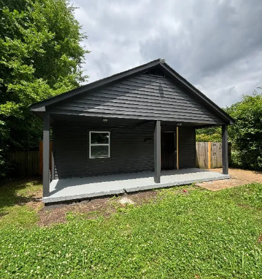 A small house with a grey roof and a white door is surrounded by greenery.