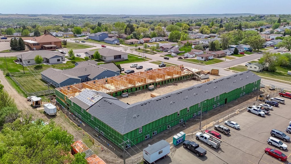 A construction site with a partially built green building in the foreground and a parking lot with cars in the background.