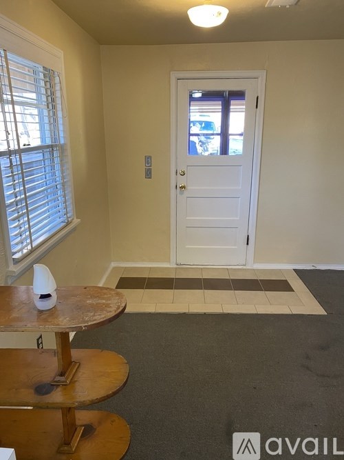 A room with a white door, a window with blinds, a wooden table with a white object on it, and a carpeted floor.