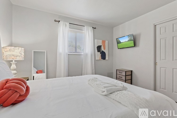 A bedroom with a large bed, a window with curtains, a lamp, a TV, and a small shelf with a picture frame.