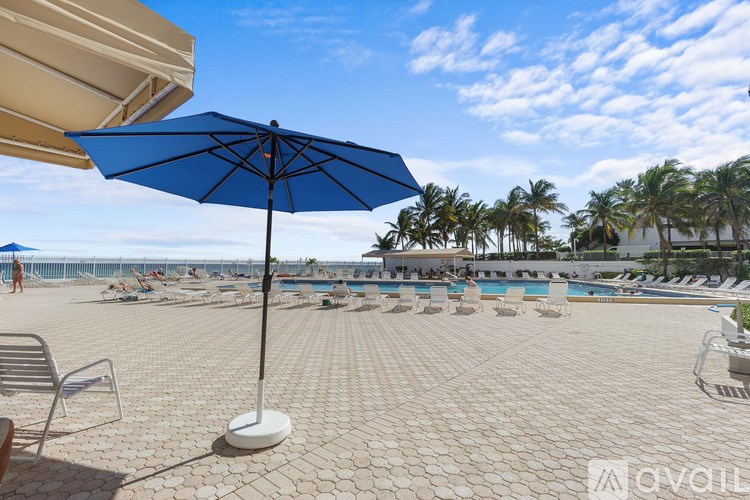 A blue umbrella stands on a patio overlooking a pool and beach.