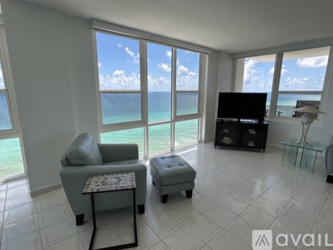 A living room with a grey sofa and ottoman, a flat screen TV, and a view of the ocean through the windows.