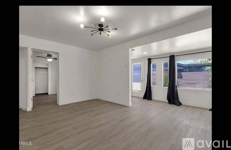 A spacious room with a ceiling fan and lighting fixture, hardwood flooring, and a window with black curtains.