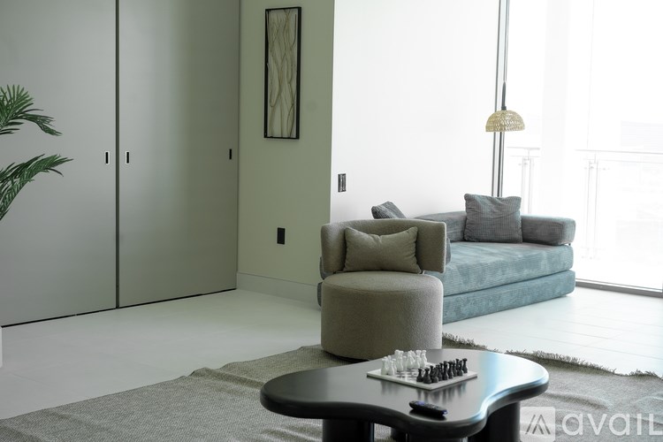 A living room with a grey armchair, a blue couch, a coffee table with a chess set on it, and a large window.