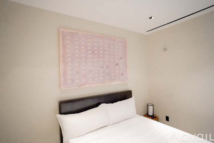 A bedroom with a bed, pillows, a nightstand, and a framed pink calendar on the wall.